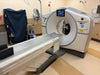 A medical imaging machine, likely a CT scanner, with a large circular gantry and a sliding table extending into the foreground, photographed in a clinical room.