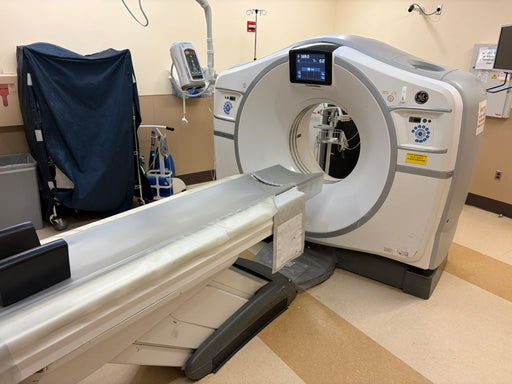 A medical imaging machine, likely a CT scanner, with a large circular gantry and a sliding table extending into the foreground, photographed in a clinical room.